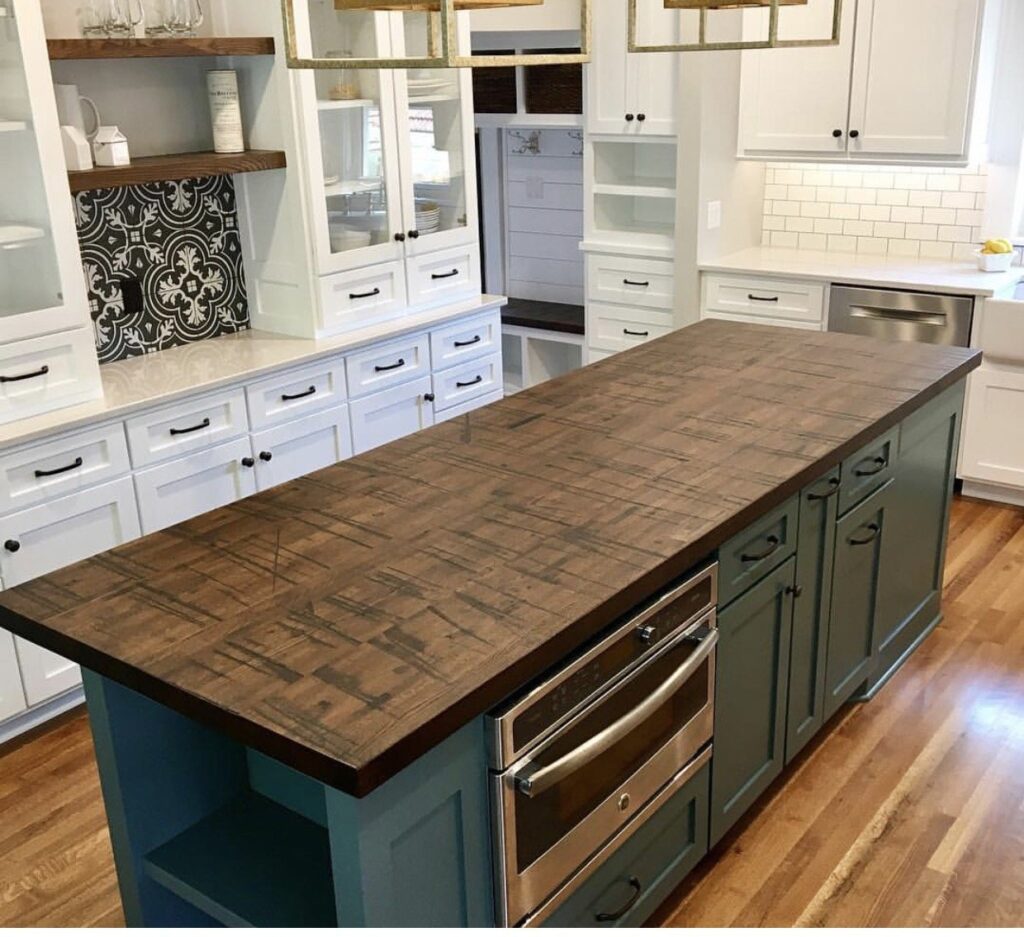 A modern kitchen with white cabinets, a wooden floor, and a central island featuring a dark wood countertop. There are built-in ovens in the island, and decorative pendant lights hang above.