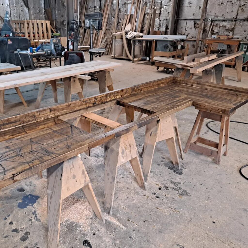 A woodworking shop with several wooden tables supported by saw horses. Wood shavings are scattered on the floor. Various tools and wooden planks are visible in the background.