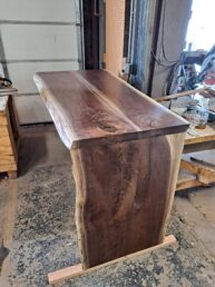 A polished wooden bar with a natural edge design is shown in a workshop setting. The wood has a rich, dark grain and is supported by a sturdy base. Workshop tools and materials are visible in the background.