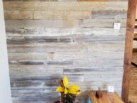 A rustic wooden wall with a small black vent at the top. Below, a colorful houseplant in a white pot stands next to a small wooden table with turquoise legs. The floor is made of reclaimed wood.