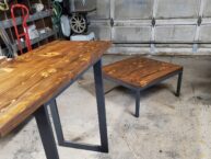 Wooden tables with metal legs on a concrete garage floor. The larger table is to the left, with a square table to the right. The garage in the background shows various tools and equipment.