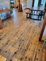 A room with wooden flooring featuring a rustic table and bench set. Plants are placed on a small table, and wood panels decorate the wall. The room has natural light coming from windows.