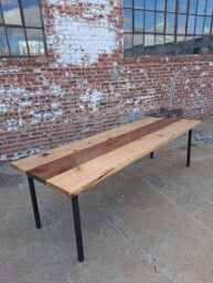 A long wooden table with metal legs stands on a concrete surface against a brick wall with several windows. The tabletop features a mix of light and dark wood tones, highlighting its natural grain.