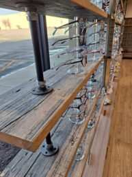 A wooden shelf with metal supports displays various eyeglasses on clear stands. The shelf is in front of a large window, revealing a parking lot and barren trees outside. The room has wooden flooring.