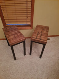 Two wooden side tables with dark metal legs are placed on a beige carpet. They are positioned at an angle, with a window with wooden blinds in the background, set against a light-colored wall.