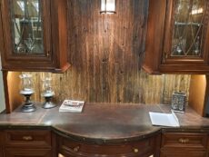 A wooden cabinet with glass-fronted storage and a curved counter displays wine bottles on lower shelves. It features decorative lighting, wooden paneling, and a small book, blending rustic and elegant design elements.