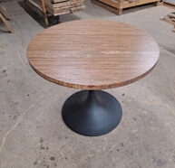 A round wooden table with a dark, pedestal base sits on a workshop floor. Stacks of wood and various tools are visible in the background, suggesting a woodworking environment.