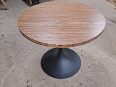 A round wooden table with a dark, pedestal base sits on a workshop floor. Stacks of wood and various tools are visible in the background, suggesting a woodworking environment.