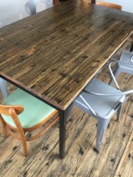 A wooden table with metal legs is surrounded by various chairs, including an upholstered wooden chair with a green cushion and several gray metal chairs. The floor has a rustic, scratched wooden appearance.