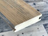 Close-up view of a wooden plank with tongue and groove joints against a wooden floor background. The planks end shows the interlocking design used for joining multiple pieces together.