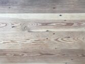 A wooden surface with visible grain patterns and natural variations in color. The wood displays a mix of light and dark tones, with some knots and lines running horizontally across the surface.