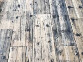 A close-up view of a distressed wooden floor with a pattern of dark stains, scratches, and wear, highlighting the rustic and weathered texture of the planks.