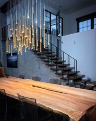 A modern dining room features a long wooden table with a natural edge, surrounded by transparent chairs. Above, numerous slender pendant lights hang in an artistic cluster. A staircase with black railings ascends in the background.