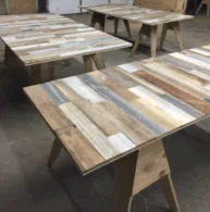 Wooden tables with plank-style surfaces and sawhorse legs are arranged in a workshop. Each table features a variety of wood tones and textures, creating a rustic and handcrafted appearance.