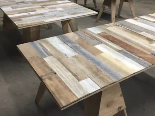 Wooden tables with plank-style surfaces and sawhorse legs are arranged in a workshop. Each table features a variety of wood tones and textures, creating a rustic and handcrafted appearance.