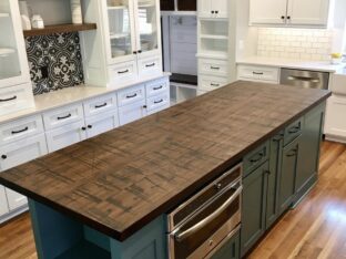 A modern kitchen with white cabinets, a wooden floor, and a central island featuring a dark wood countertop. There are built-in ovens in the island, and decorative pendant lights hang above.