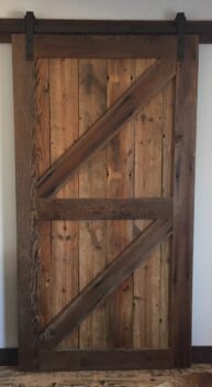 A rustic wooden barn door with a diagonal Z-brace design, made of weathered, vertically aligned wooden planks. It hangs on a black metal sliding track above a wooden floor. The door exudes a vintage, farmhouse aesthetic.