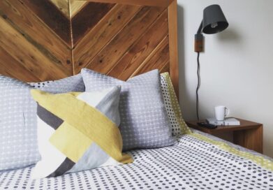 A cozy bedroom with a wooden headboard, geometric-patterned bedding in gray and yellow tones, and a matching pillow. A black wall-mounted lamp is above a wooden nightstand, which holds a white mug and a small notepad with a pen.