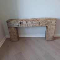 Rough-hewn wooden mantelpiece with a natural, rustic look, placed against a plain white wall on light wood flooring. An electrical outlet is visible on the wall behind it.