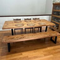 A wooden dining table set with six intricately designed wicker placemats, accompanied by a matching bench and three wooden chairs, all set on a polished hardwood floor. A shelf with glassware is visible on the right.