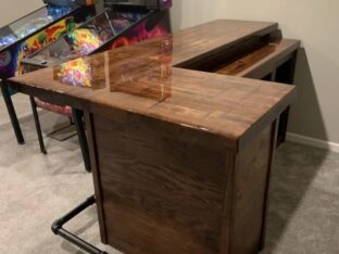 A polished wooden bar with a corner design stands on a carpeted floor, adjacent to vintage pinball machines with colorful designs. A metal footrest is attached to the bottom of the bar.