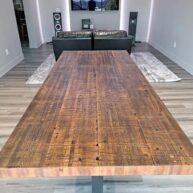 A long wooden dining table with a rustic finish in a modern living room. The room features wood flooring, two large speakers, a TV on the wall, and a leather sofa set on a textured rug.