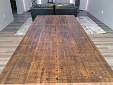 A long wooden dining table with a rustic finish in a modern living room. The room features wood flooring, two large speakers, a TV on the wall, and a leather sofa set on a textured rug.