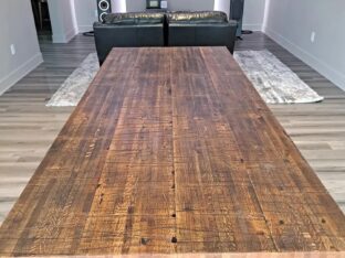 A long wooden dining table with a rustic finish in a modern living room. The room features wood flooring, two large speakers, a TV on the wall, and a leather sofa set on a textured rug.