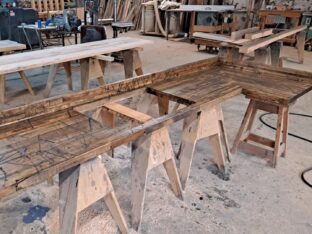 A woodworking shop with several wooden tables supported by saw horses. Wood shavings are scattered on the floor. Various tools and wooden planks are visible in the background.