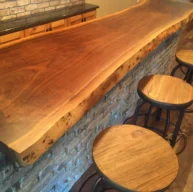 A Live Edge Tops wooden bar counter with a natural finish is set against a brick wall. Four round wooden stools with metal frames are neatly aligned along the bar, which sits atop a floor made of wood planks.