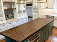 A modern kitchen with a long wooden island countertop. The island has teal cabinets and stainless steel appliances. Surrounding white cabinetry features open shelves, a black and white decorative backsplash, and a tile backsplash near the sink.