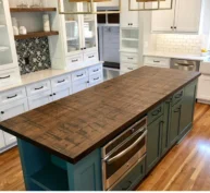 A modern kitchen with a long wooden island countertop. The island has teal cabinets and stainless steel appliances. Surrounding white cabinetry features open shelves, a black and white decorative backsplash, and a tile backsplash near the sink.