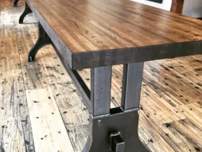 A long dining table crafted from a dark reclaimed rail car plank, featuring sturdy black metal legs on a rustic wooden floor, situated in a room with light walls and various wall samples in the background.