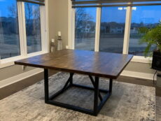 A dining room with a square wooden table on a patterned rug. Large windows provide a view of the outside, and a modern chandelier hangs above the table. The room features neutral colors and a potted plant in the corner.