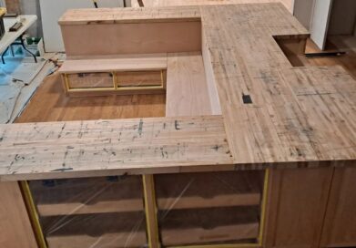 A kitchen under renovation with bare wooden cabinets and countertops. Construction materials and tools are scattered around. A ladder leans against the wall, and the floor is unfinished wood.