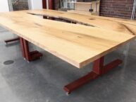 A large wooden table with a natural finish sits on red metal legs in a modern room with a concrete floor and brick walls. The tabletop features unique grain patterns and a central split design.