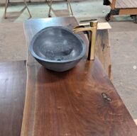 A live-edge wooden countertop with a smooth black stone vessel sink and a sleek gold faucet. The countertop showcases natural wood grain and texture, creating a rustic yet modern aesthetic.