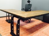 A modern office setup with a large L-shaped wooden desk on a red patterned rug. The desk has black metal supports and an ergonomic black chair with a headrest is placed behind it. The background wall is gray.