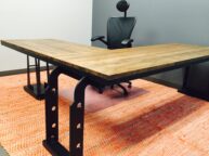 A modern office setup with a large L-shaped wooden desk on a red patterned rug. The desk has black metal supports and an ergonomic black chair with a headrest is placed behind it. The background wall is gray.