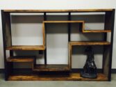 A wooden, geometrically designed bookcase with staggered shelves stands against a white wall. A small black statue of a cowboy is placed on the bottom right shelf, adding a decorative touch to the minimalist furniture piece.