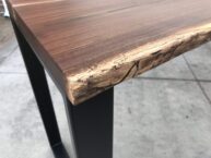 A close-up of a wooden table corner with a natural, live edge finish. The tabletop is smooth with visible wood grain patterns, and it rests on sturdy black metal legs. The background is a concrete floor.