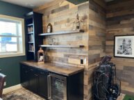 Home office with rustic wooden walls and shelves displaying books and decor. A wooden countertop has a mini-fridge below. The framed picture on the wall and a window with blinds complete the cozy setting.